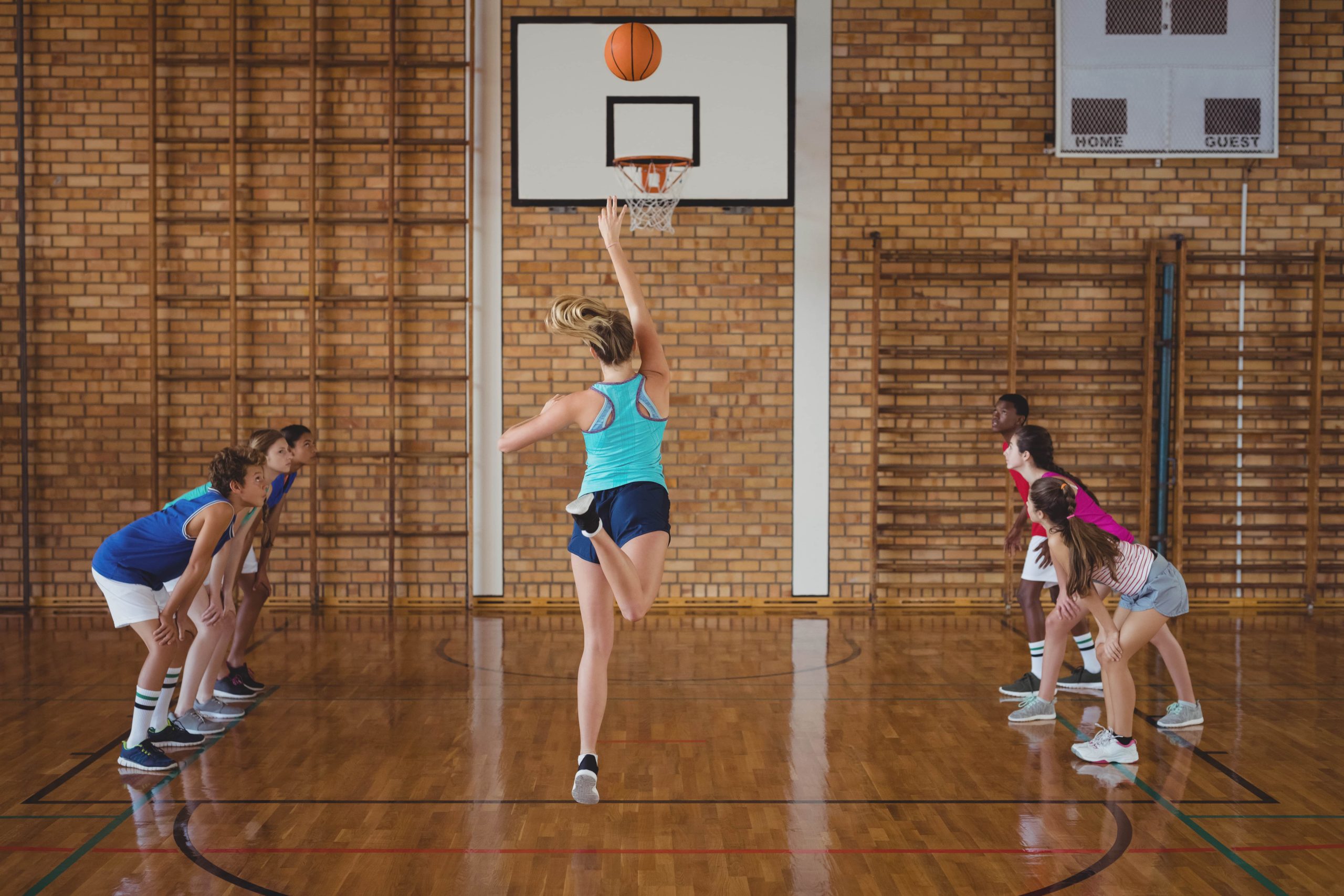 Imagen de estudiantes jugando baloncesto en un gimnasio escolar, mostrando trabajo en equipo y actividad física